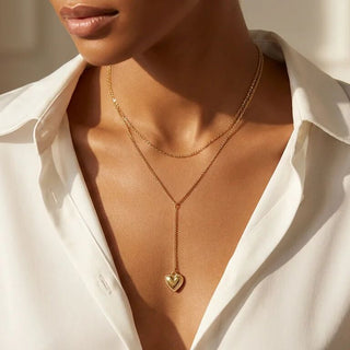 Close-up of a person wearing a gold necklace with a heart pendant, against a neutral background.