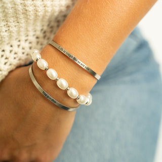 Two bracelets on a wrist with pearls and engraved text, against a blurred background.