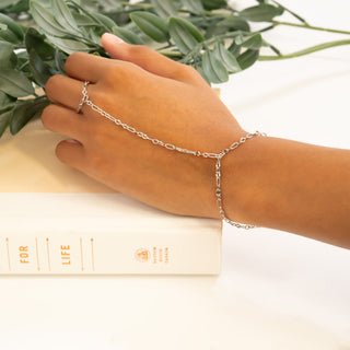 Hand wearing a silver hand chain with a plant and book in the background