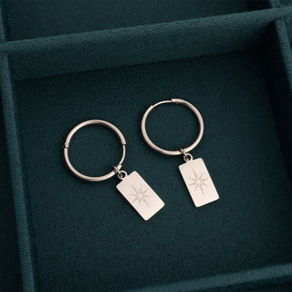 Pair of silver hoop earrings with engraved tags on a dark green fabric background