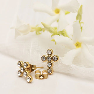 Gold cross-shaped earrings with clear stones on a white fabric background with flowers.