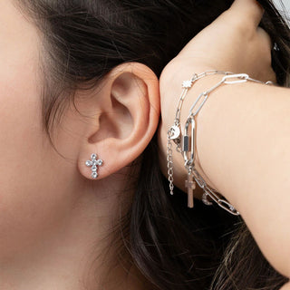Close-up of an ear with a silver cross earring and a hand wearing multiple silver bracelets.