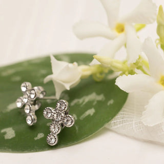 Silver cross-shaped earrings on a green leaf with white flowers in the background