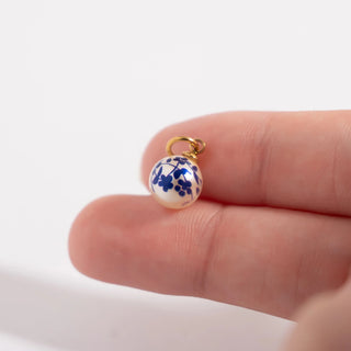 Small blue and white ceramic charm on a gold chain held between fingers against a white background