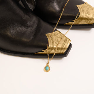 Gold necklace with a turquoise pendant draped over black leather cowboy boots on a white background