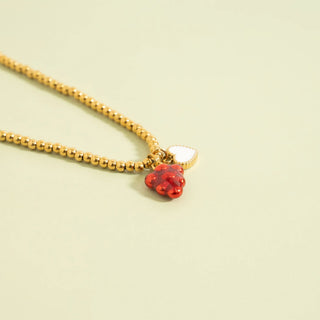 Gold beaded necklace with a red berry and white heart-shaped pendant on a light green background