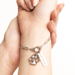 Hand wearing a silver bracelet with paw charm and 'LET'S GO' tag on a white background