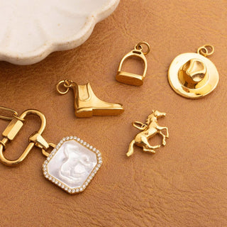 Collection of gold equestrian charms including a boot, stirrup, horse, and carabiner necklace on a brown surface.