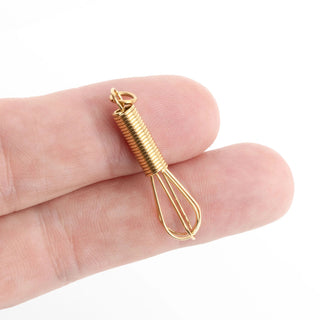 Gold whisk charm held between fingers on a white background