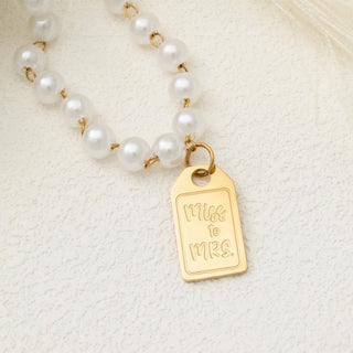 Pearl necklace with a gold 'Miss to Mrs.' tag on a textured beige background