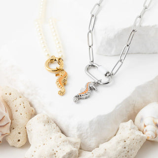 Two seahorse-themed necklaces on a white surface with shells and sand.