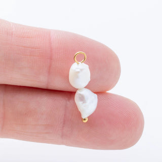 Two pearl charms on gold loops held between fingers against a white background