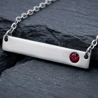 Silver necklace with a horizontal bar pendant featuring a red gemstone on a dark stone surface.